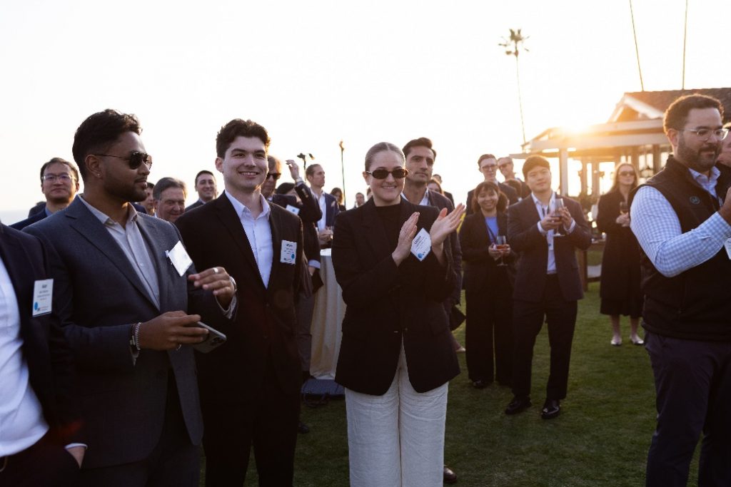 A crowd on a lawn claps, with the sun setting behind them.